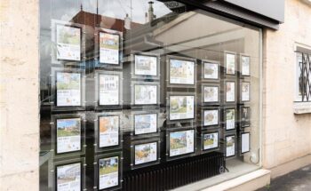 Agent immobilier présentant un contrat de vente à un couple d’acheteurs dans une agence — explication des frais d’agence immobilière à l’achat, qui paie et quand.